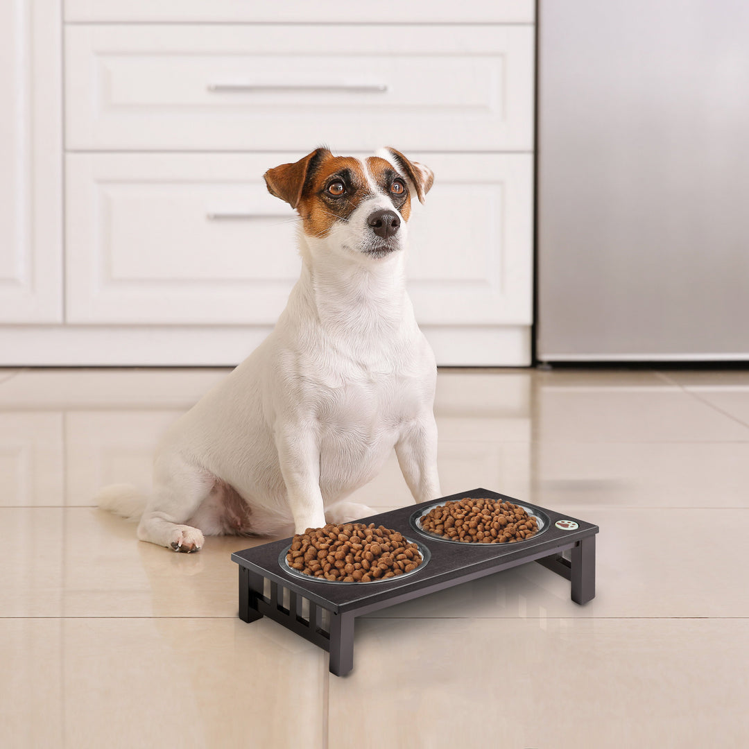 A small white and brown dog at his black feeder with 2 Stainless Steel Bowls