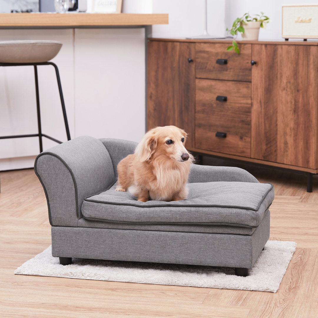 A small brown dog sits on a gray dog-sized chaise lounge next to a brown sideboard.