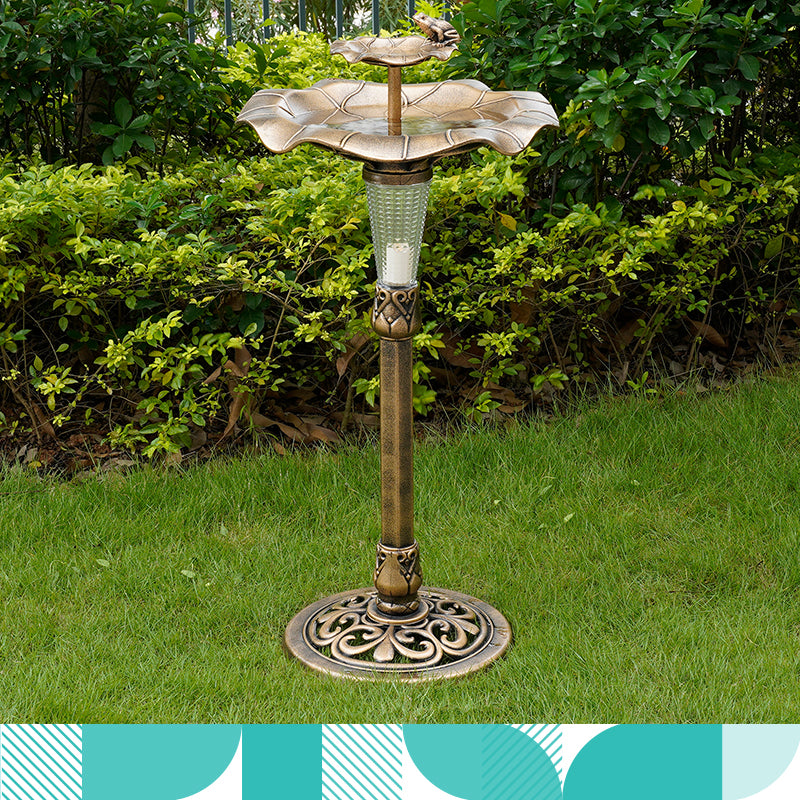 A brass birdbath with build in solar powered light sits in the grass next to a garden.