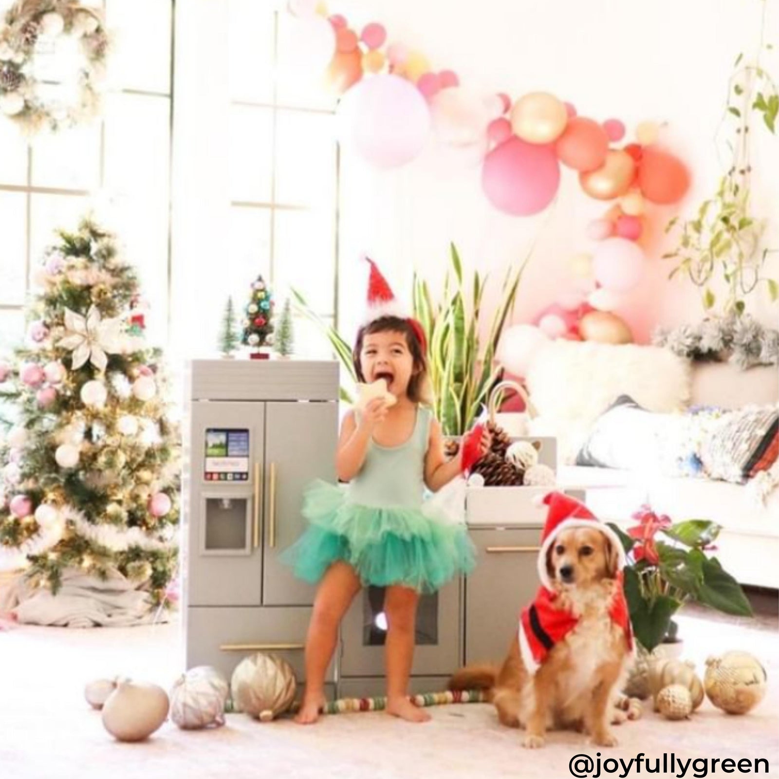 A girl and a dog wearing Christmas outfit standing in front of a grey play kitchen