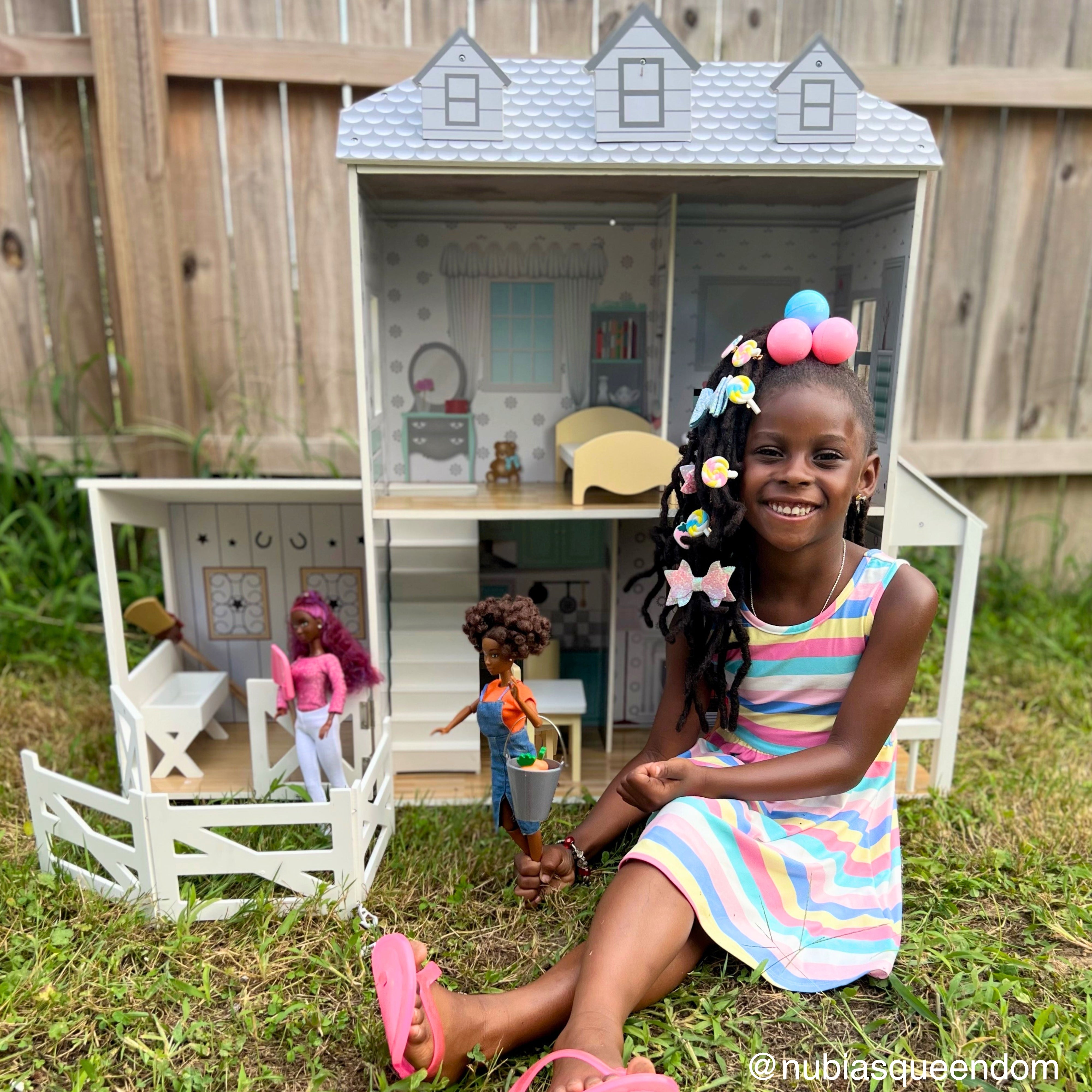 a girl sits proudly in front of her farmhouse style dollhouse with her fashion dolls