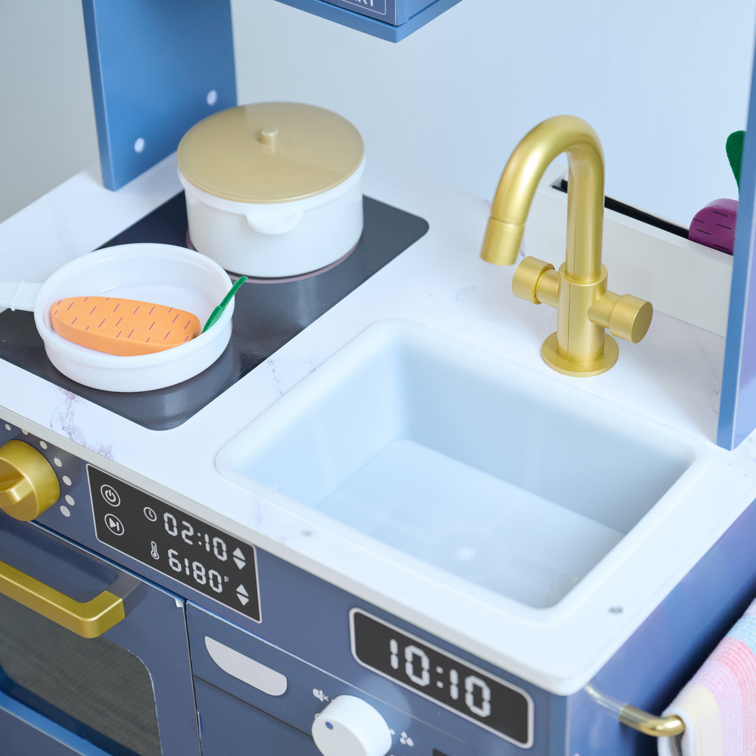 Toy kitchen set with stovetop, gold faucet, sink, and play food including a wooden carrot in a frying pan and a white pot.