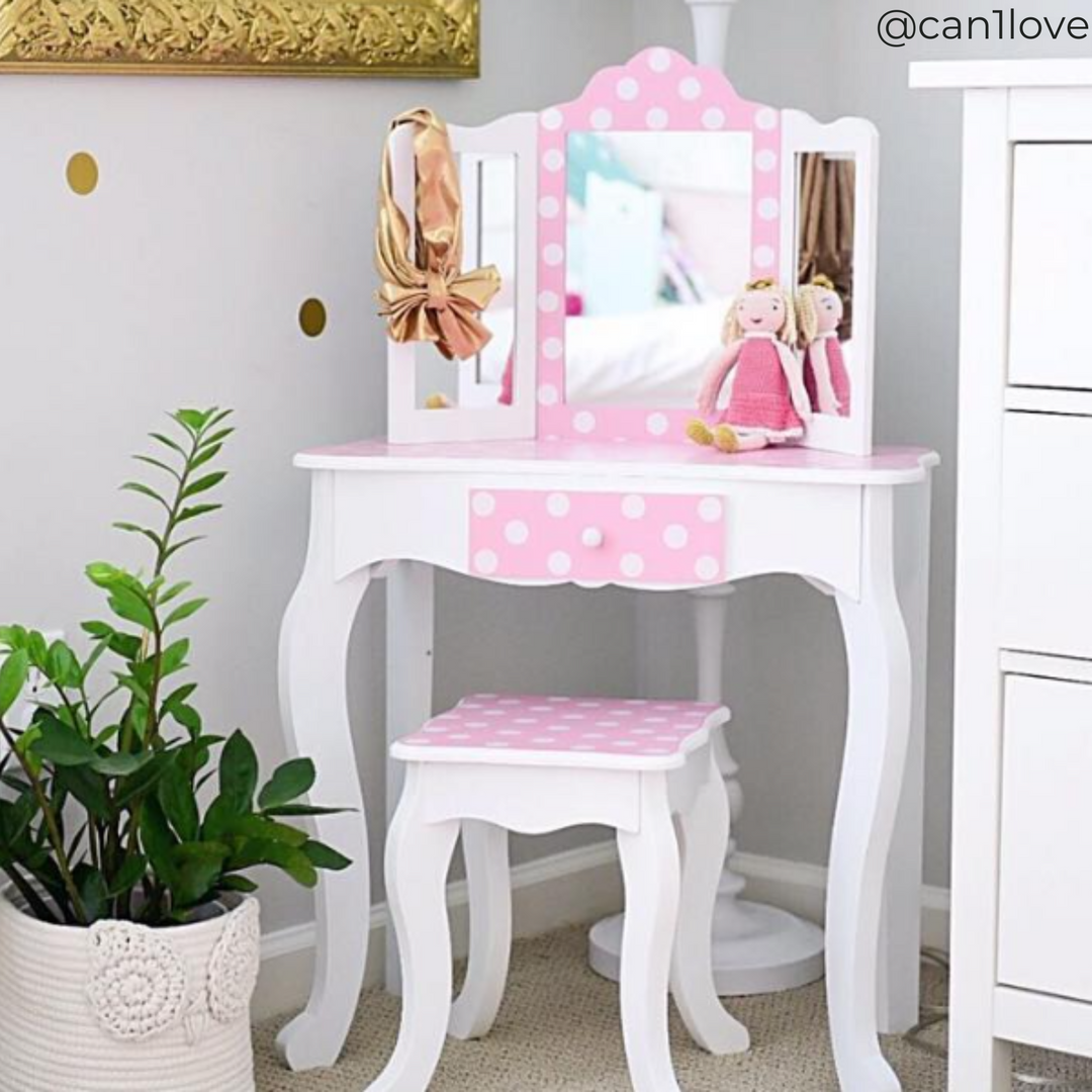 A kids' vanity table and matching stool with trifold mirror, white with pink and white polka dot accents in a bedroom with a doll and hair bow on the table