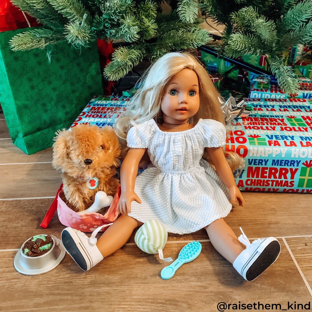 An 18" blonde doll in a gingham dress with a stuffed brown dog and dog accessories under a Christmas tree