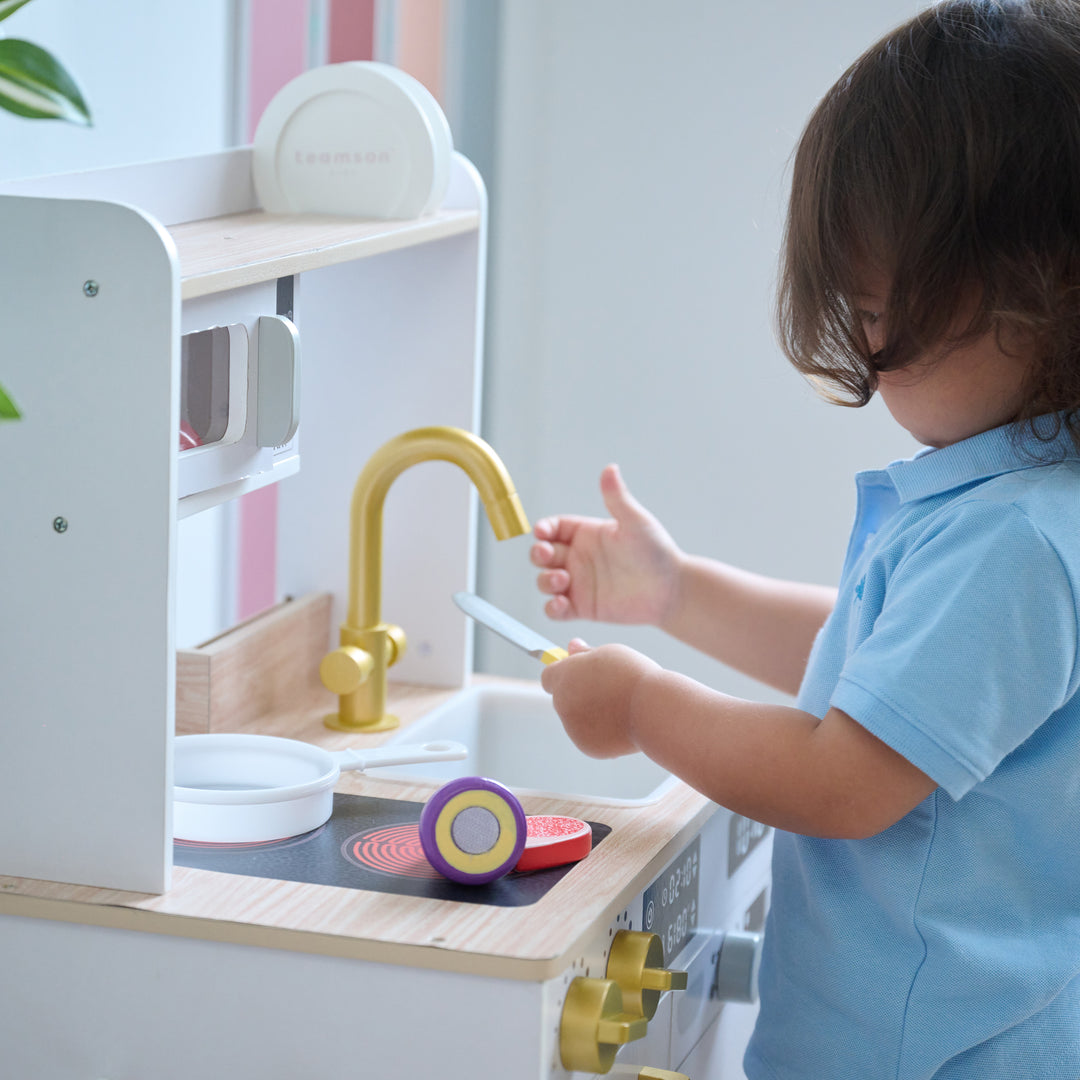 Child playing with a wooden toy knife and food at a wooden play kitchen, with gold faucet, stovetop, and microwave visible.