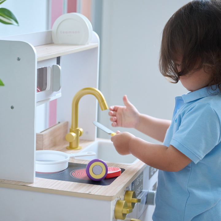 Child playing with a wooden toy knife and food at a wooden play kitchen, with gold faucet, stovetop, and microwave visible.