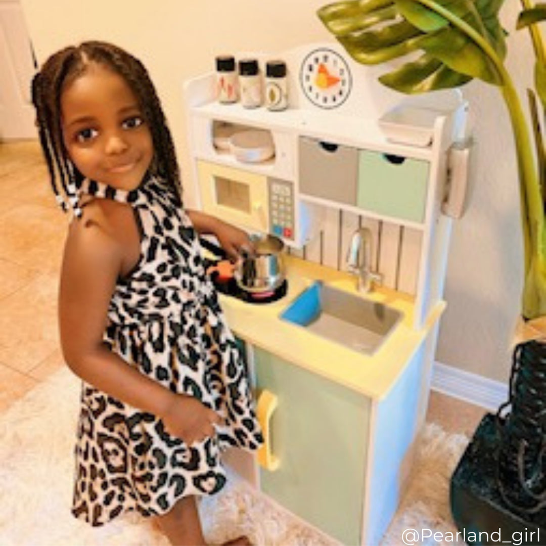 A little girl standing a play kitchen with her hand on a pretend pot