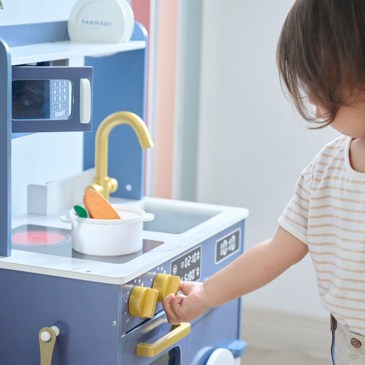 Child playing with blue wooden play kitchen, turning knobs while a pot with toy carrot sits on the stovetop next to a gold faucet.