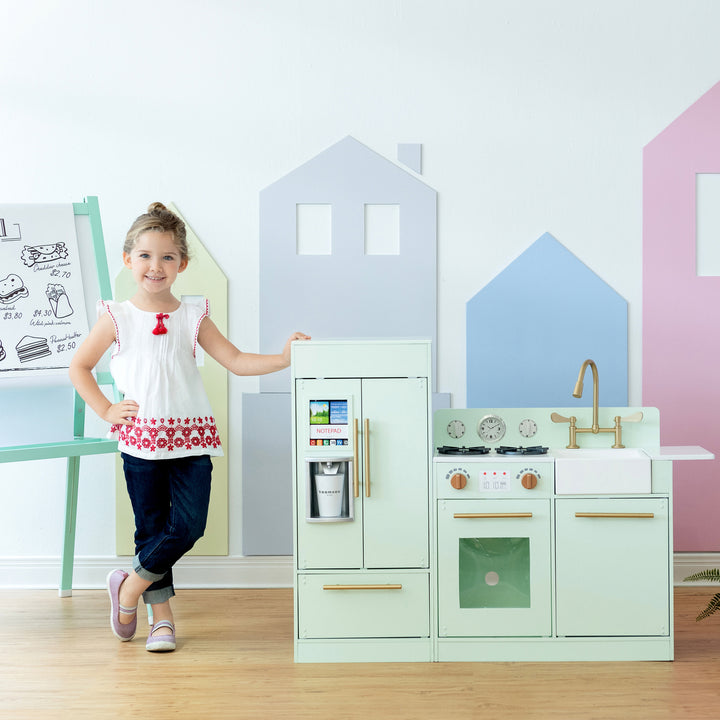 A girl standing by a modern-style mint play kitchen with gold hardware