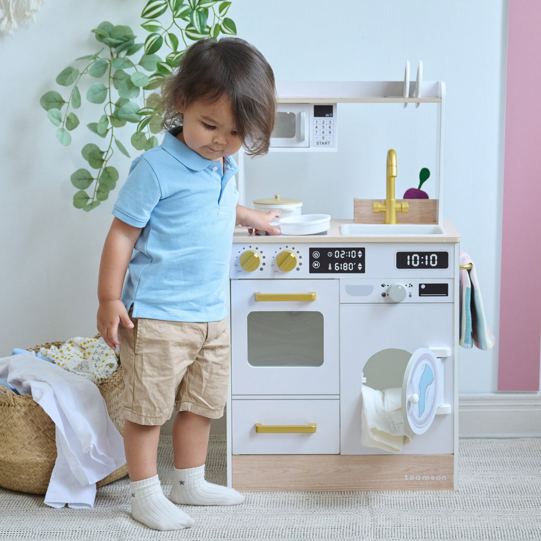 Child standing beside a wooden play kitchen, putting a pan on the stovetop, washer doors open and toy dishes on the counter.