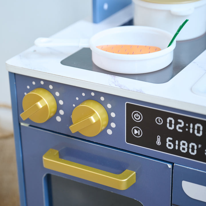 Close-up of toy kitchen stovetop with gold knobs, digital display, and a frying pan cooking a wooden carrot