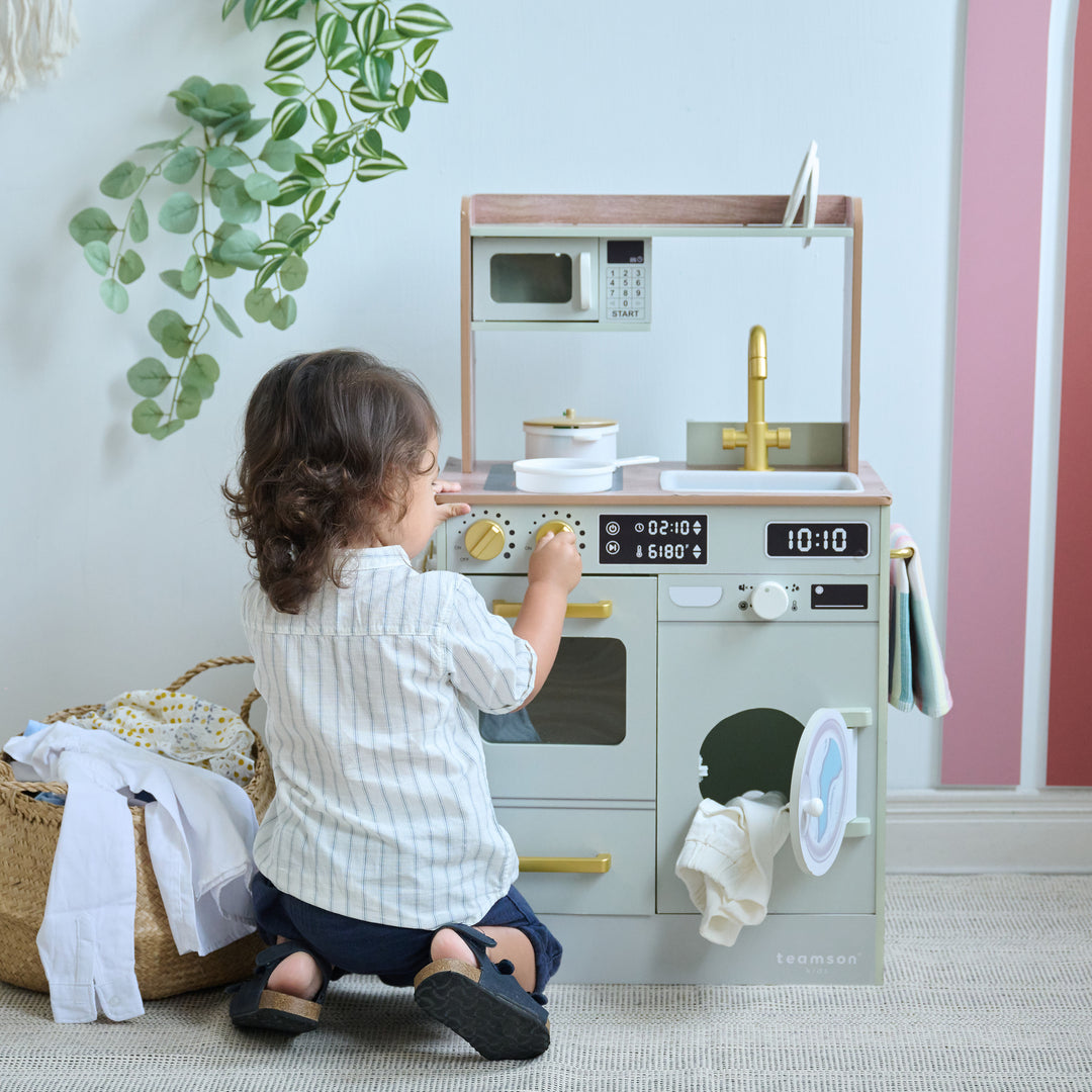 Child kneeling to turn knobs on a kids kitchen-laundry playset with toy oven, sink, microwave, and washer.