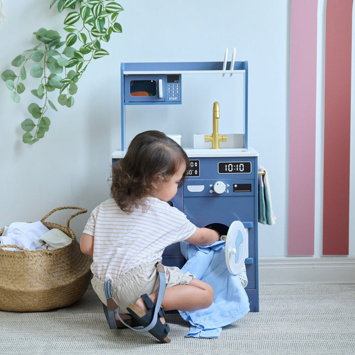 Child loading play laundry into the washing machine section of a blue kitchen-laundry combo playset.