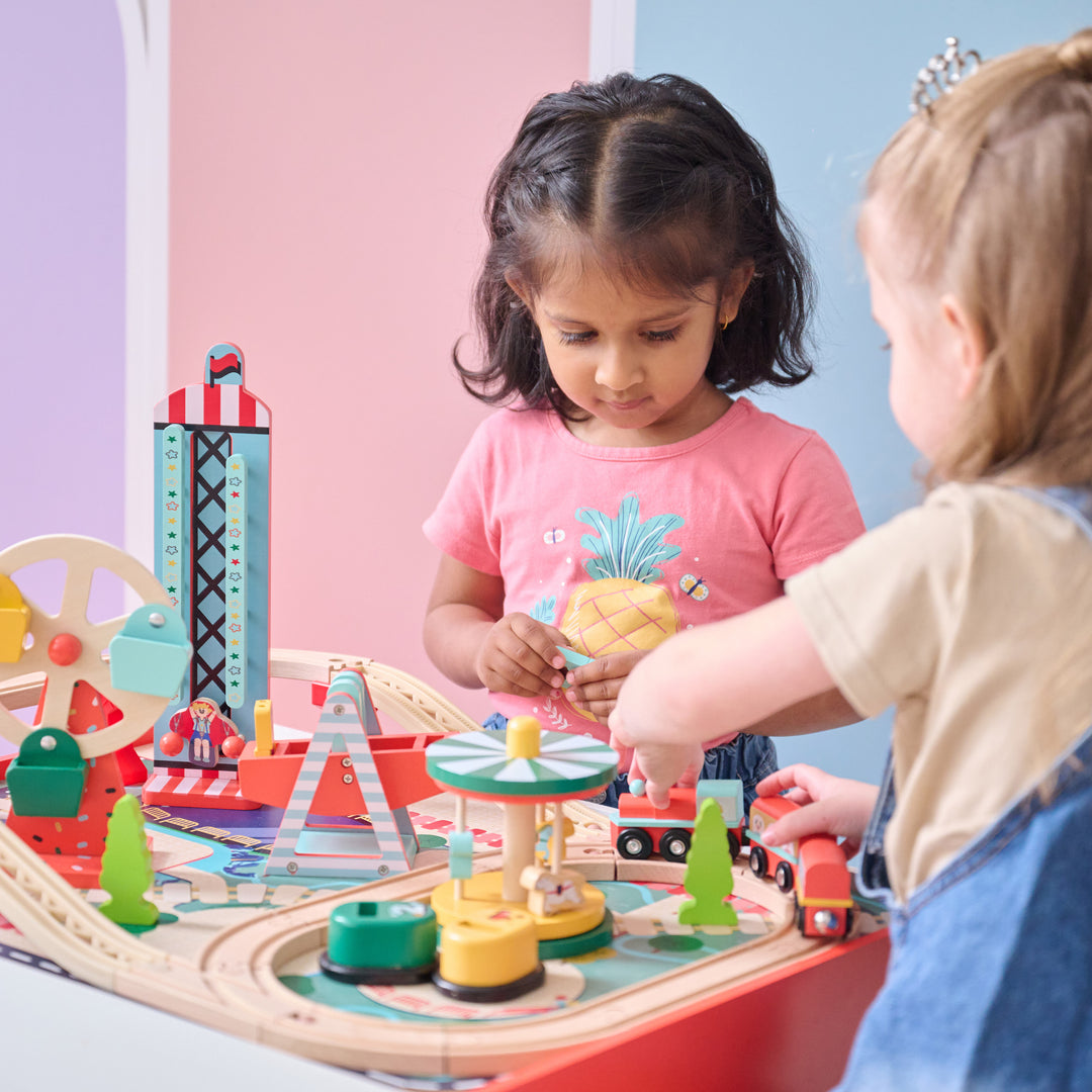 Two kids playing with a wooden amusement park train table filled with colorful tracks, rides, and figures.