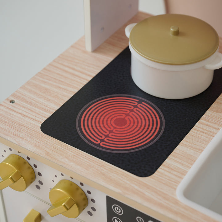 Close-up of toy stovetop with red burner graphic, gold knobs, and a white pot with lid on a wooden play kitchen set.