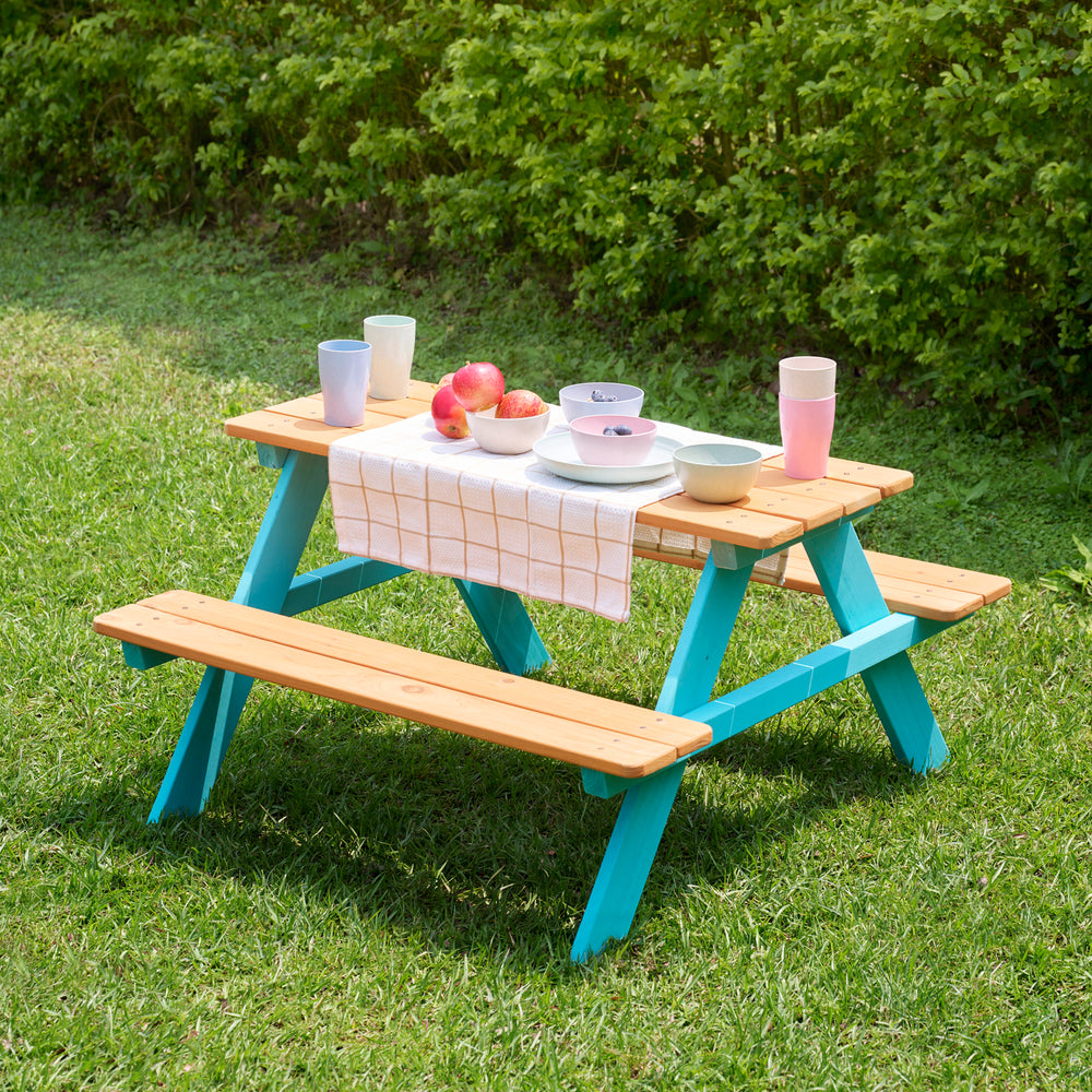 Teamson Kids Child Sized Wooden Outdoor Picnic Table in a sunny park, with picnic setting on the table