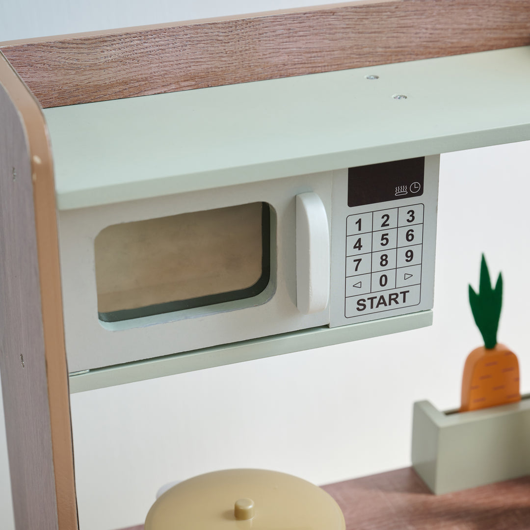 Close-up of toy microwave with keypad and handle above a play stovetop, next to a mini garden with a wooden carrot.