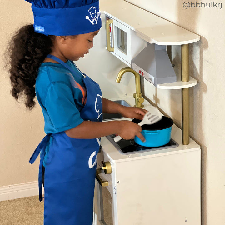 A little girl dressed in a chef's uniform pretending to cook on her play kitchen