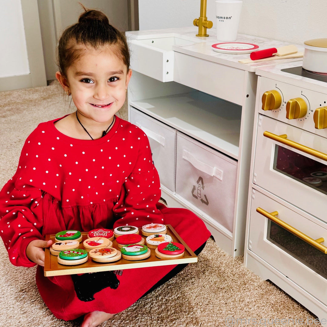 A little girl with a tray of pretend christmas cookies next to a play kitchen