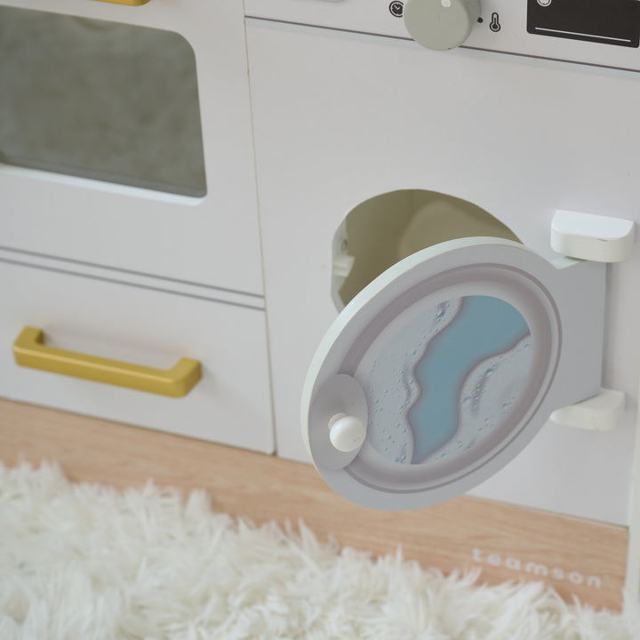 Close-up of toy washing machine with door open, showing interior drum and printed water design, next to a toy oven on a play kitchen.