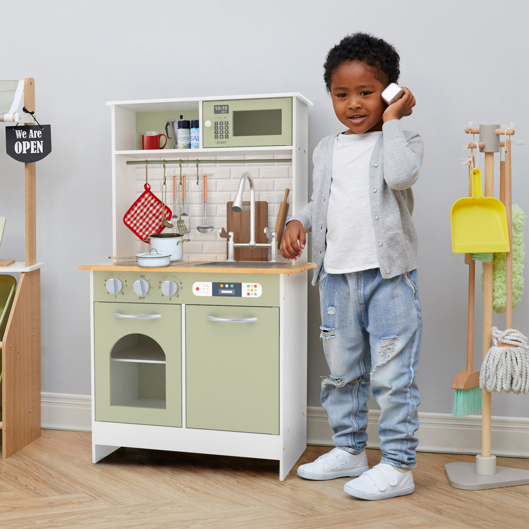 A little boy talking on the pretend telephone next to a play kitchen.