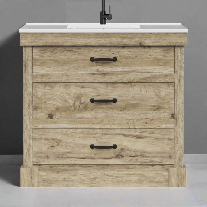 Light oak finished sink vanity with white ceramic countertop and basin and black hardware against a light gray wall.