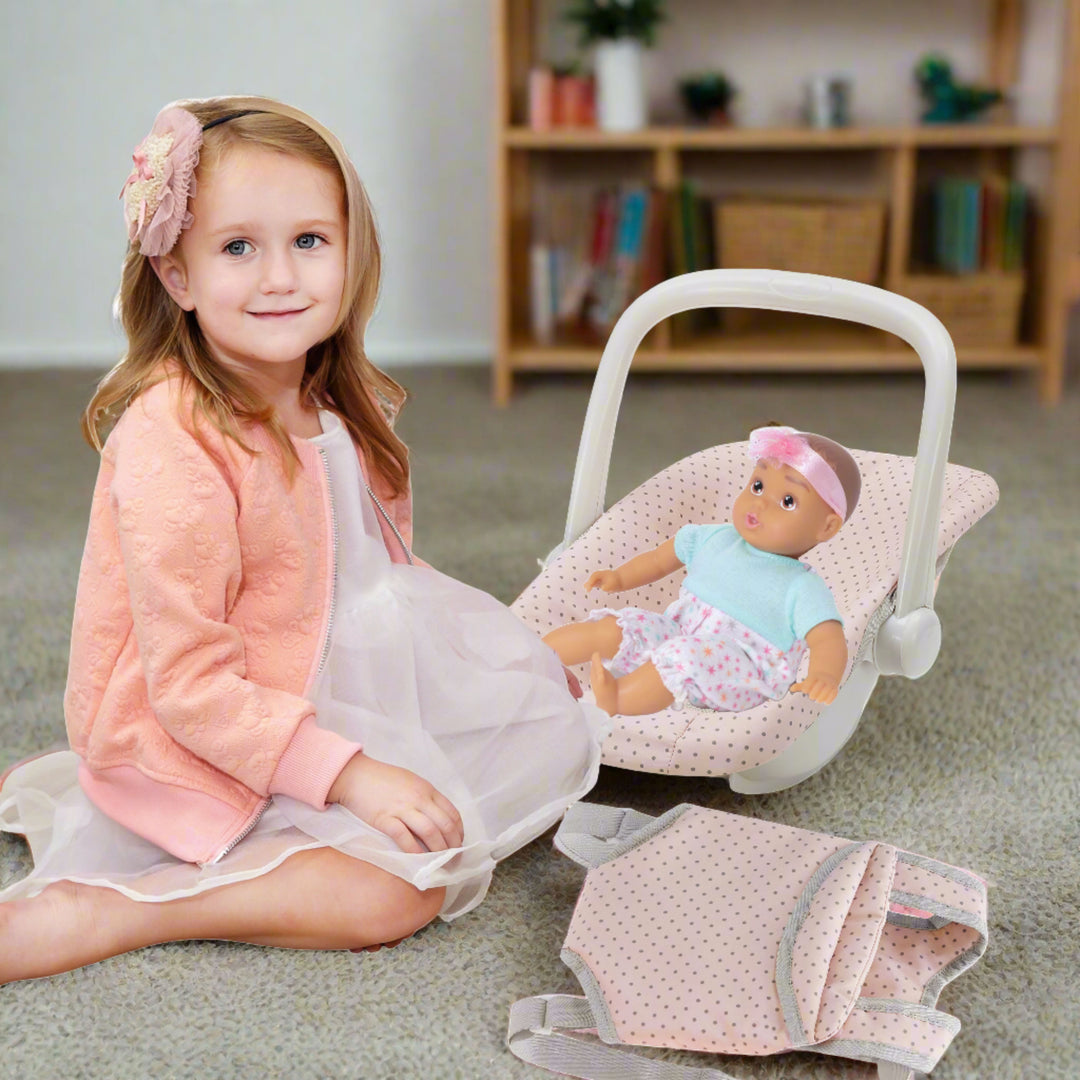 young girl sitting next to her doll in car seat carrier, in a living room