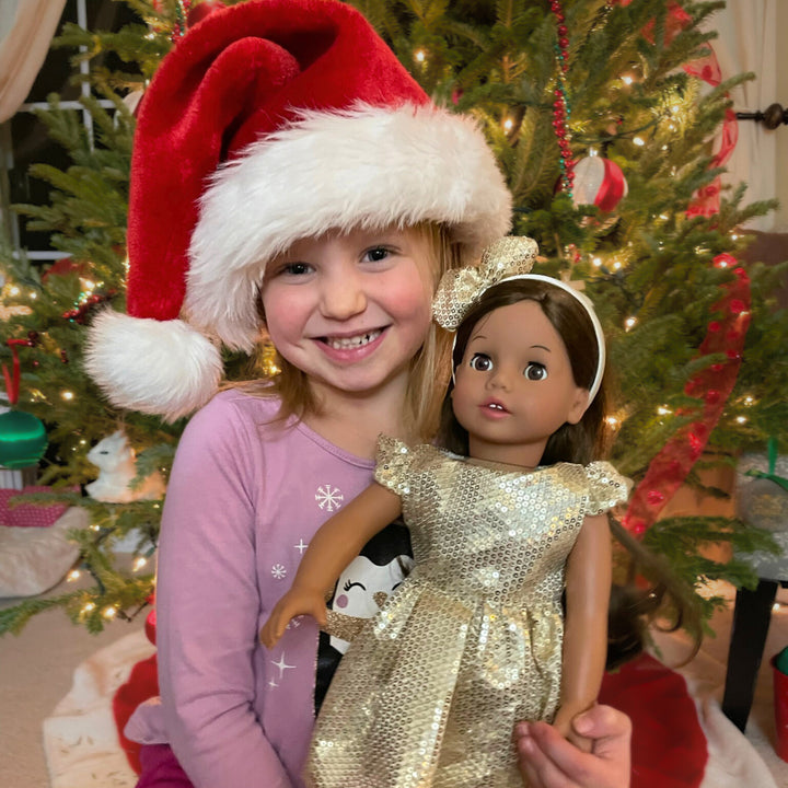 A little girl in a Santa hat holding a brunette doll in a sparkly dress and headband