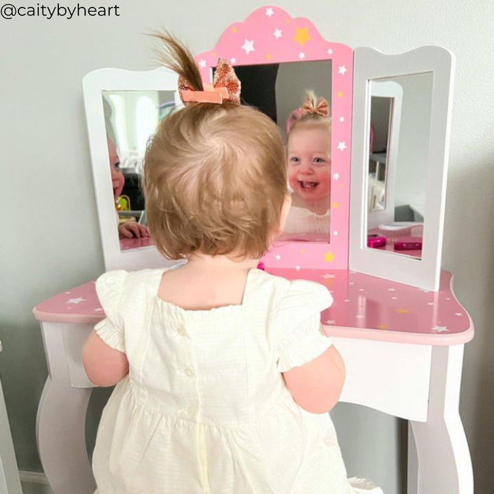 A little girl sitting at a white vanity with pink accents looking into the mirror