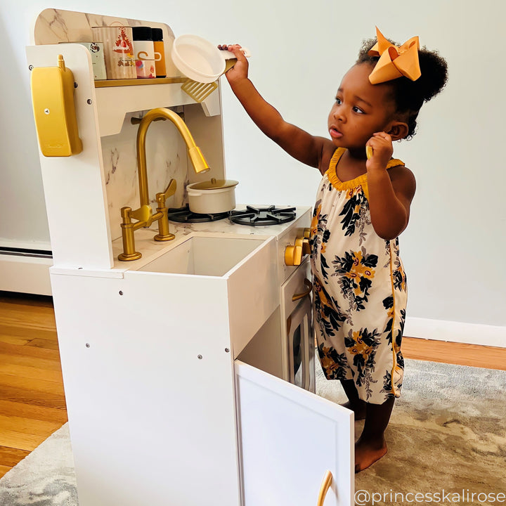 A little girl putting a white pan up on a shelf on a white and gold play kitchen
