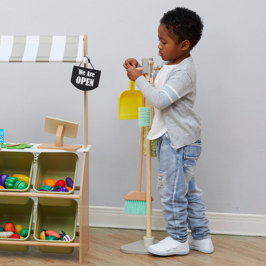 A little boy takes a yellow dust pan off a organizational stand for a play cleaning set