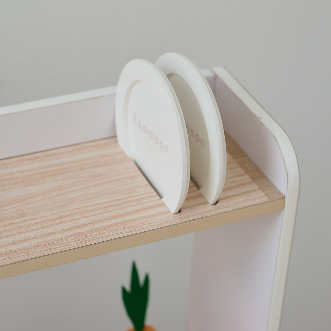 Two white toy plates stored upright on a dish drainer on the top shelf above a mini garden, part of a children's play kitchen.