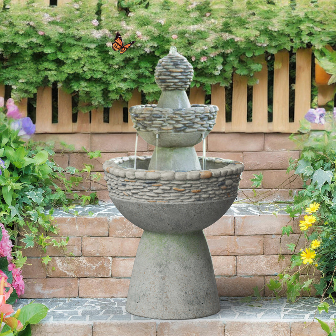 A 3-tiered birdbath style water fountain with pebble accents in front of a brick and wood wall