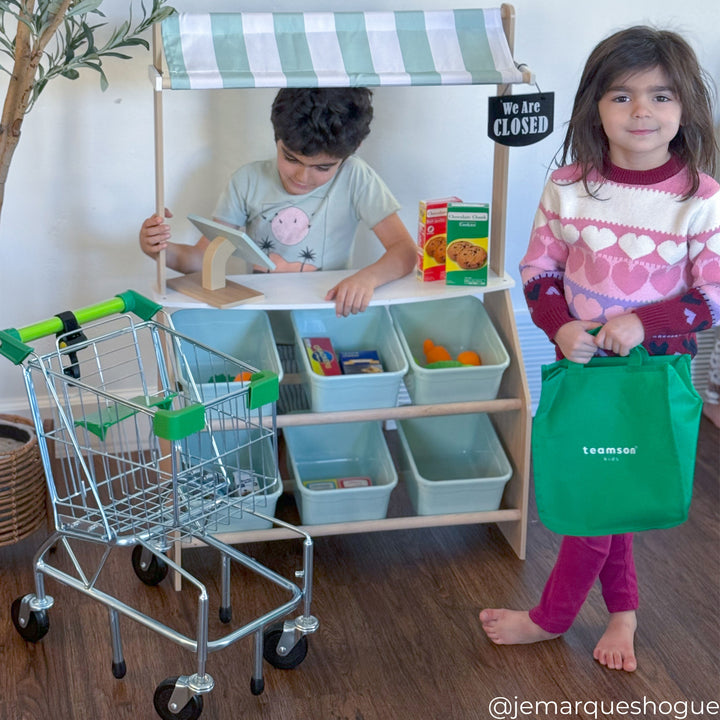 A boy standing behind a child's market stand and a girl standing with a green grocery bag