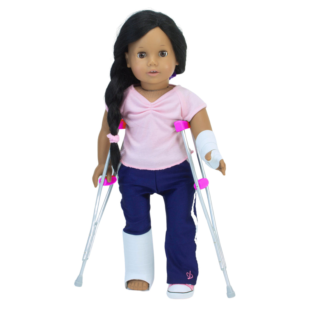 A brunette 18" Sophia's doll in navy pants and a pink tee with a wrist bandage and leg cast, balancing on pretend crutches.