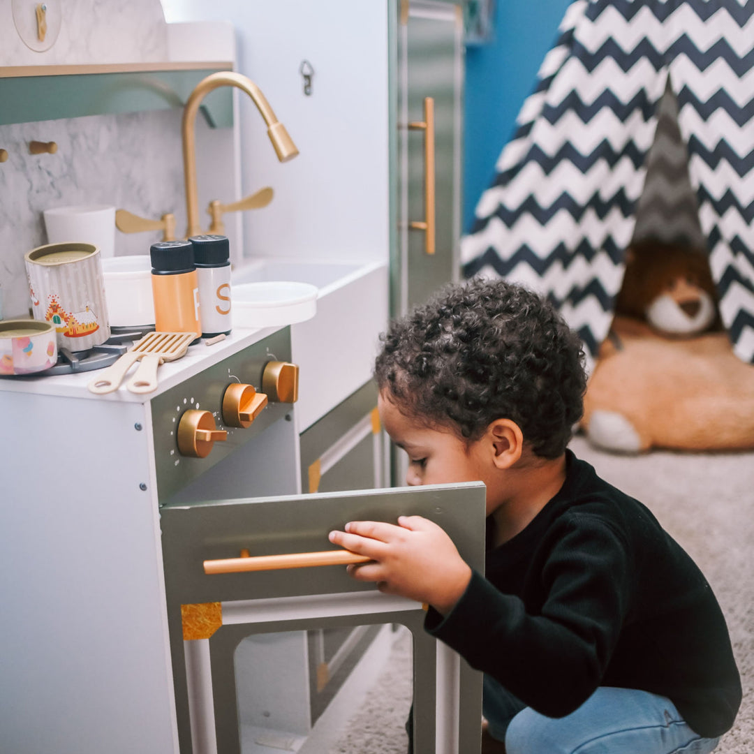 Child playing with a green and white toy kitchen set, opening the oven door, with a chevron-patterned play tent in the background.