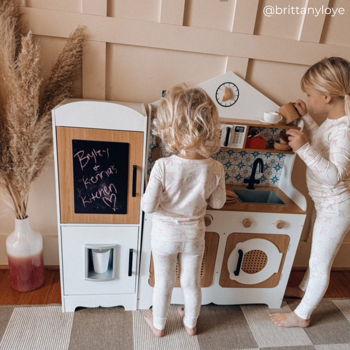 Two children in pajamas play with a toy kitchen set. One child stands by the stovetop, and the other pretends to fill a cup of tea.