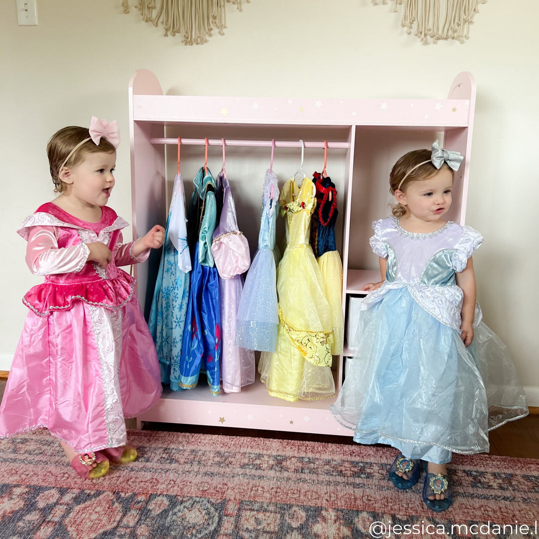 Two little girls dressed in princess costumes stand next to their clothing rack with other princess dresses hanging inside it.